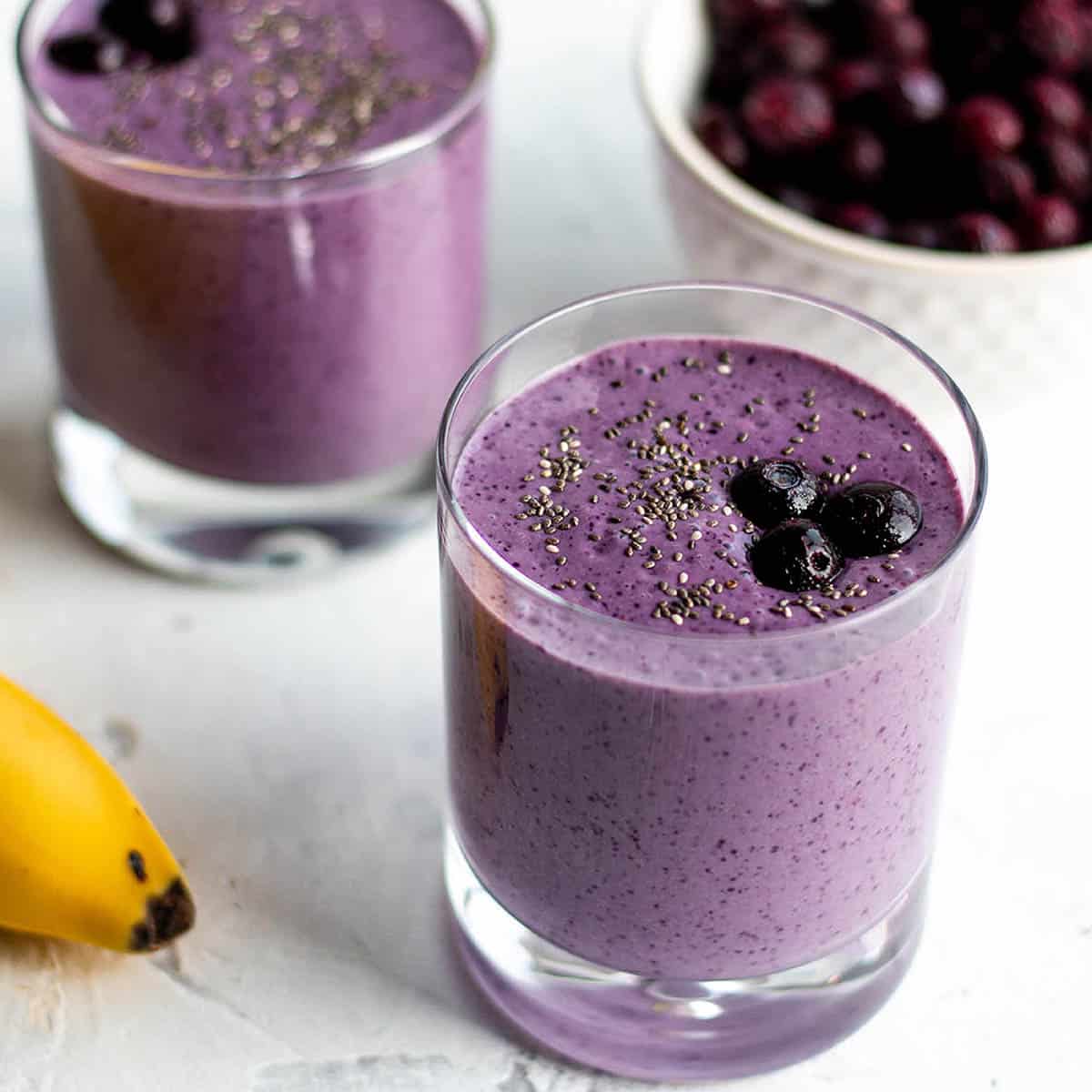 Two small glasses with a purple smoothie topped with blueberries with a bowl of blueberry in the back and a banana.