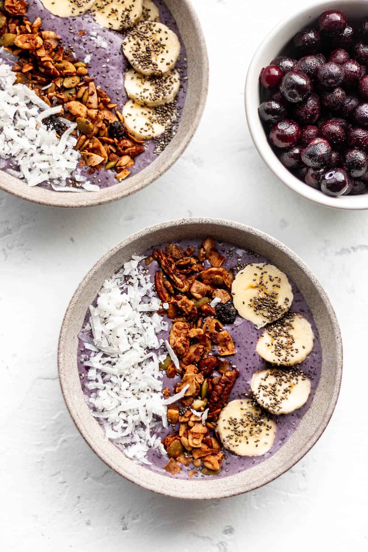 Two bowls with a purple smoothie topped with granola, banana, chai seeds and shredded coconut.

