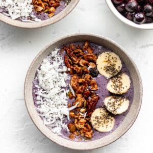 A bowl with a purple smoothie toped with coconut, granola, and banana slices.