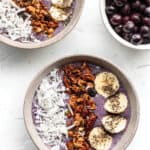 Two bowls with a purple smoothie topped with granola, banana, chai seeds and coconut.