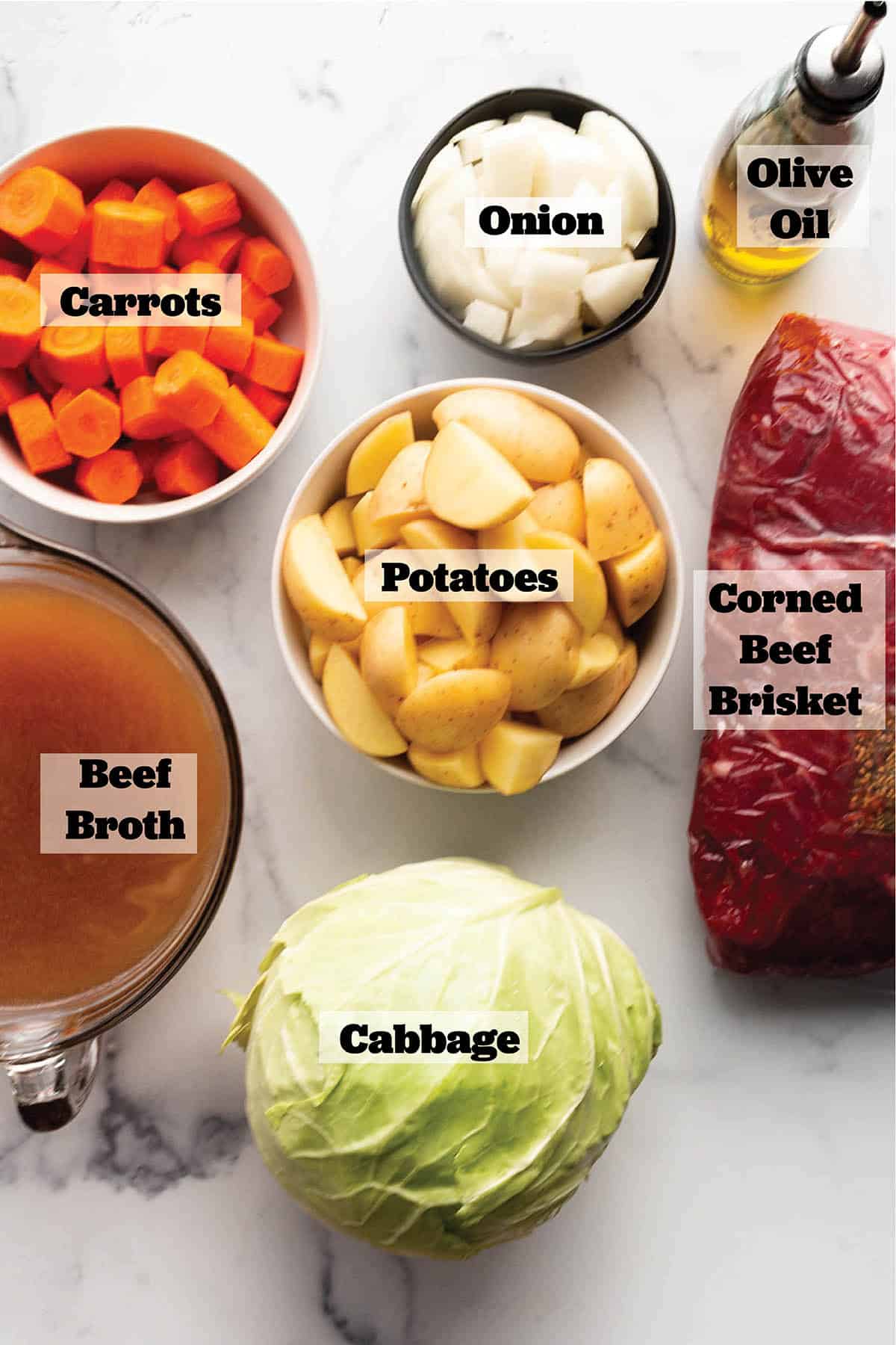 Prep bowls with chopped carrots, onions, potatoes, a head of cabbage, bowl of beef broth and a corned beef brisket and olive oil.