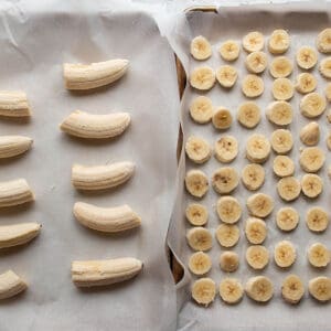 Two sheet pans lined with parchment paper, one with sliced bananas and one with halved bananas.