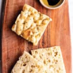 A piece of focaccia and one piece sliced open on a wooden board with a dish of olive oil and vinegerette.