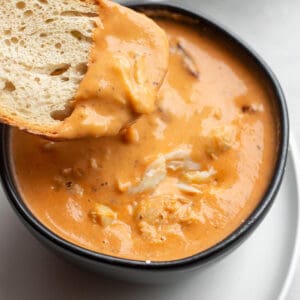 A black bowl with a crab bisque and piece of sourdough being dunked in it.