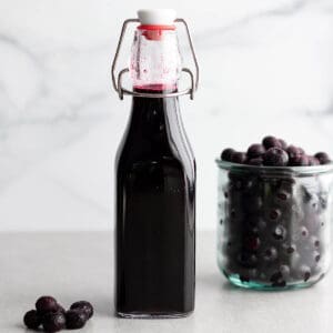 A glass bottle with a blue syrup liquid with a cup of fresh blueberries.