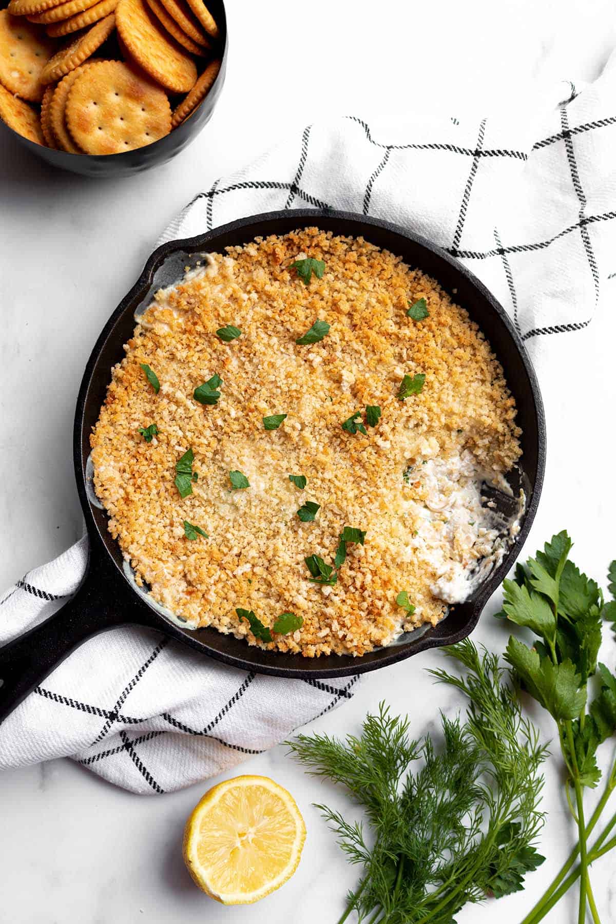A cast iron pan with a dip topped with panko crumbs with a lemon, fresh herbs and a bowl of crackers. 