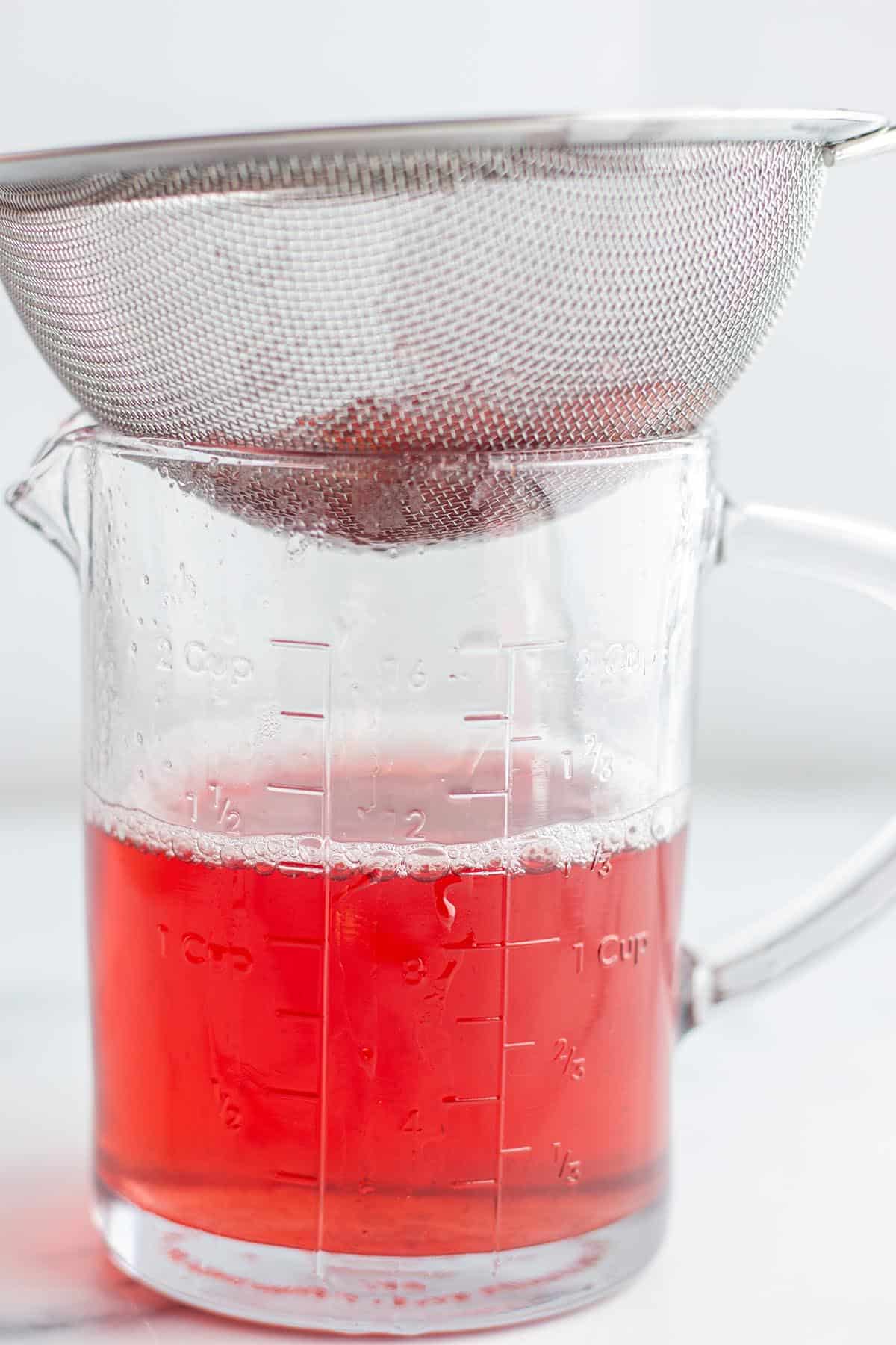 A liquid measuring cup with a pink liquid inside and a fine mesh strainer sitting on top.