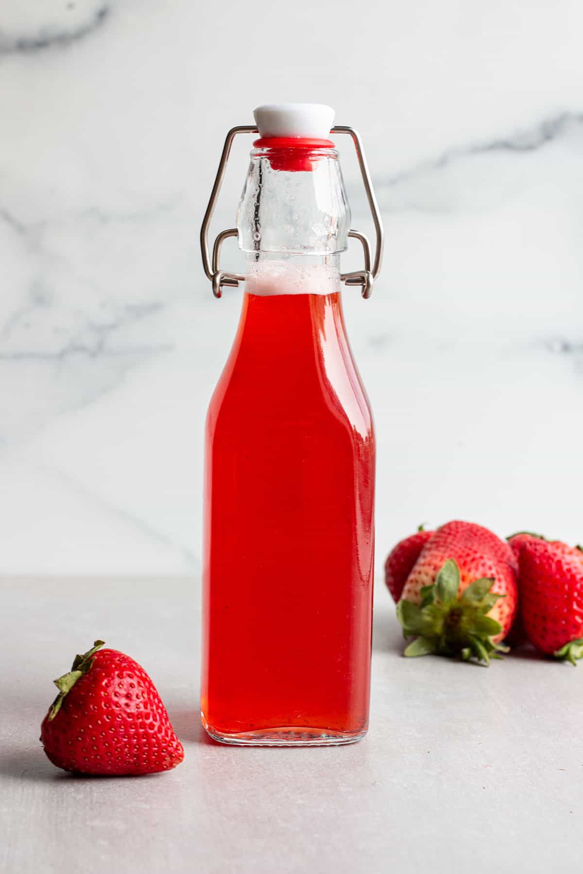 A glass flip lock bottle with a red strawberry simple syrup and fresh strawberries.