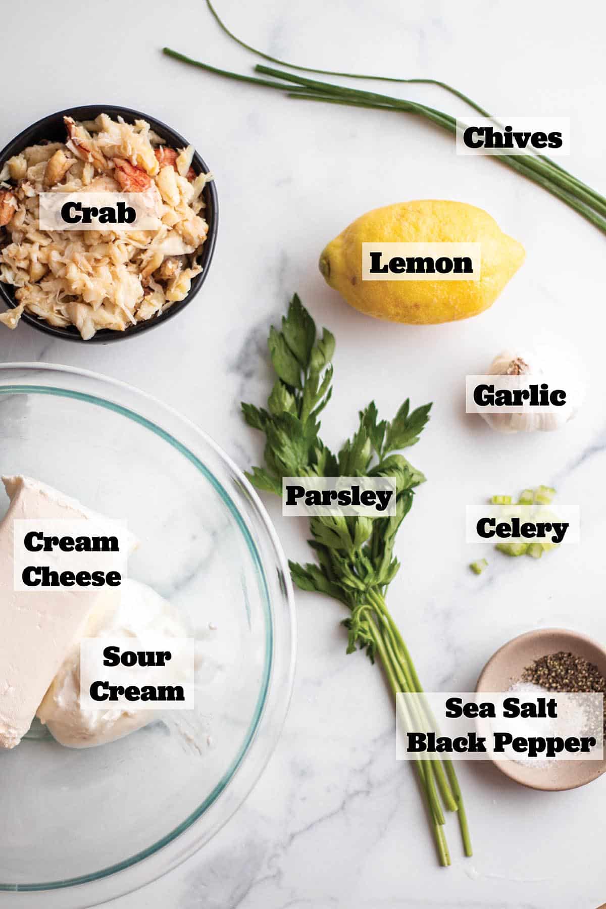 Ingredients to make a crab dip. A bowl of fresh crab, fresh chives and parsley, cream cheese, sour cream, salt & pepper, garlic, celery and lemon.