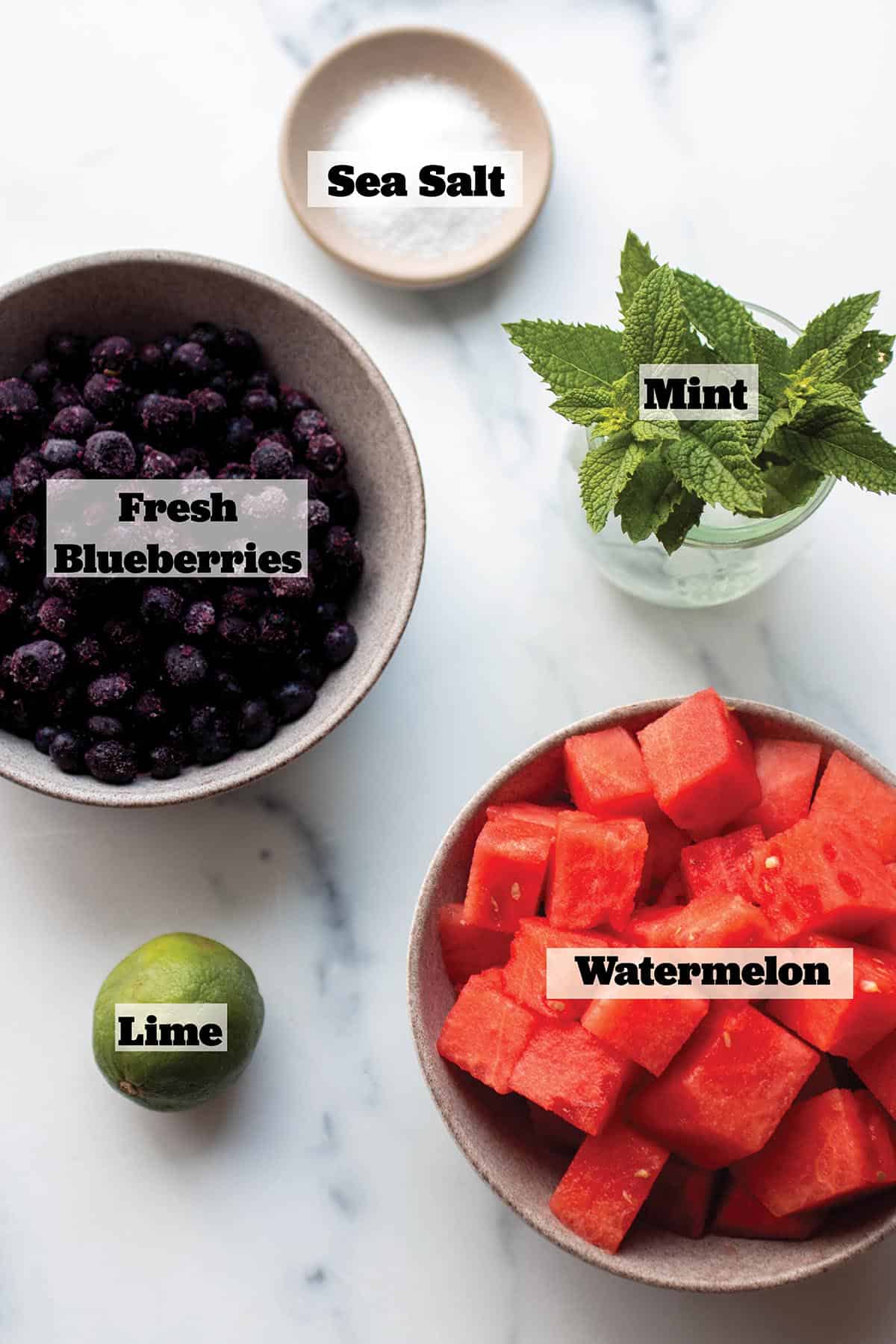 A bowl of diced watermelon, bowl of blueberries, fresh mint, a lime and dish of sea salt.