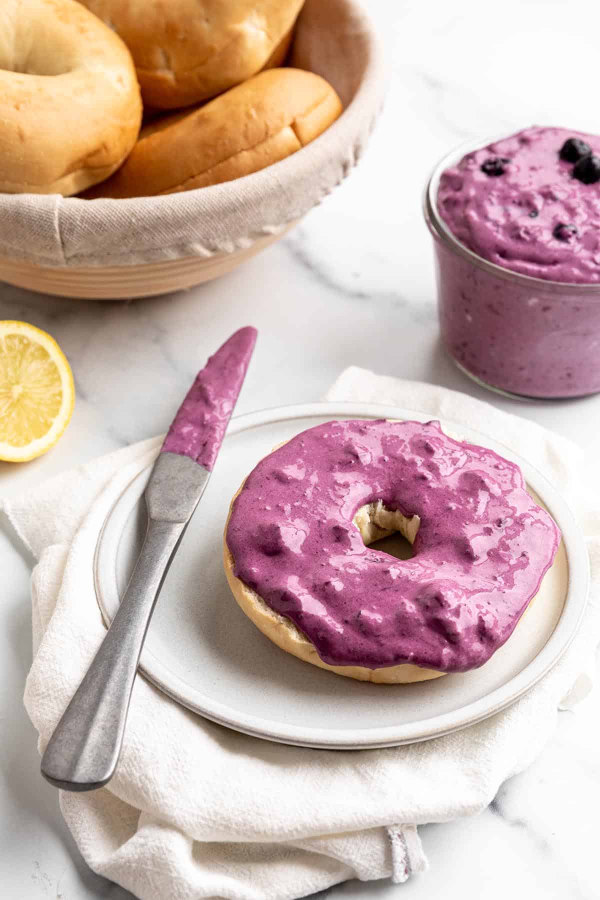 A bagel with a blueberry cream cheese spread, basket of bagels and a fresh lemon. 