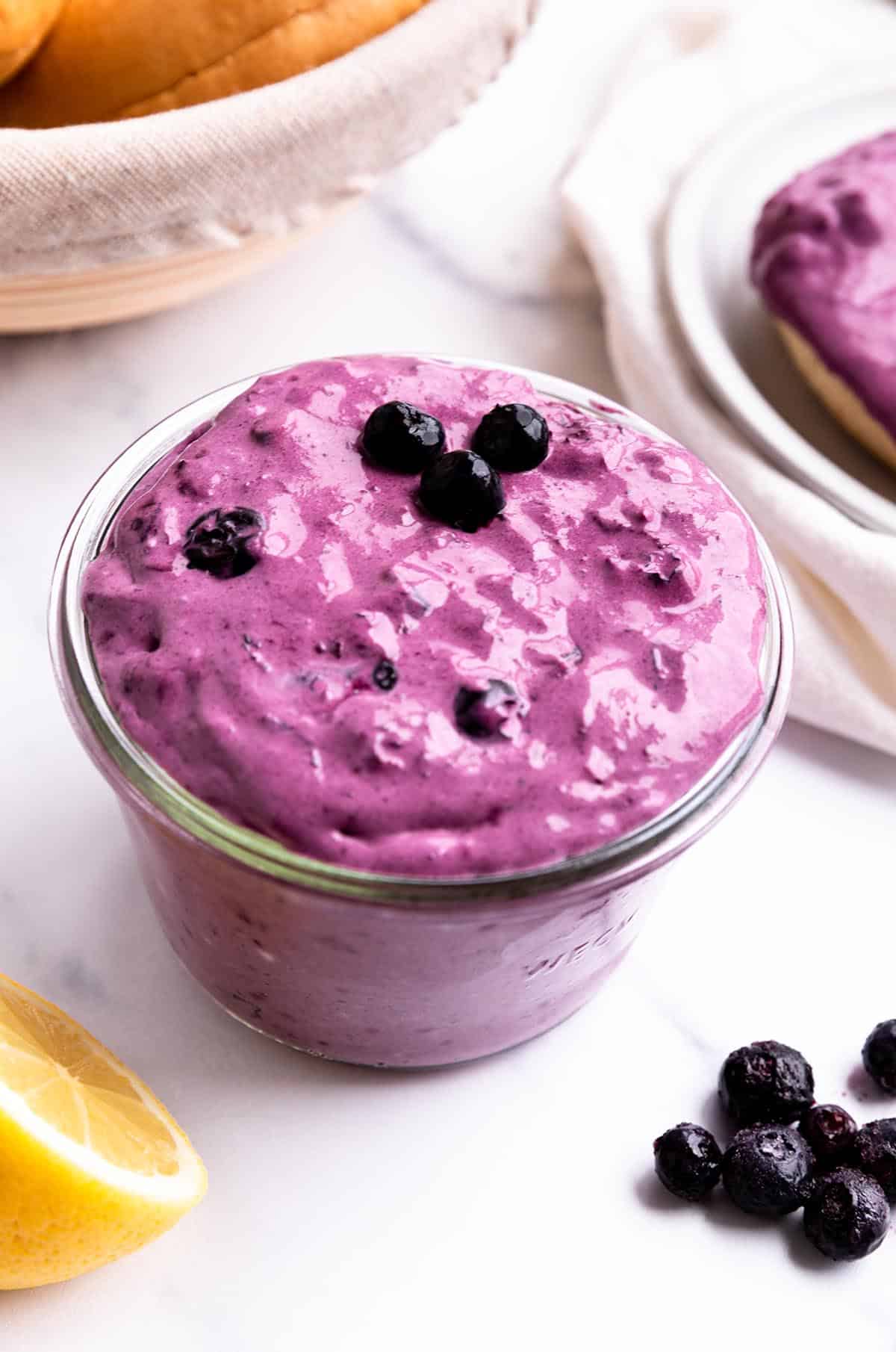A jar of blueberry cream cheese with a fresh lemon and basket of bagels. 