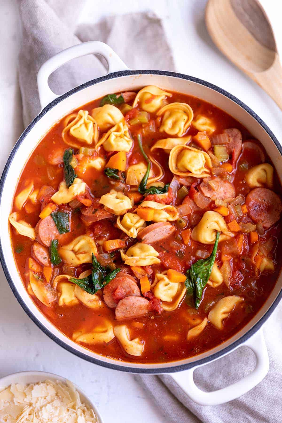 A white Dutch Oven pot with a soup full of sausage, tortellini and spinach.