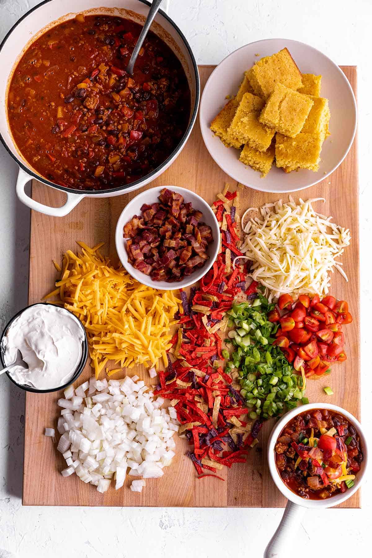 A wooden board with a pot of chili and different toppings like cheese, sour cream, bacon and green onions. 